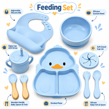 A baby feeding set arranged on a white background with callout labels. The set includes a light blue silicone suction bowl, adjustable silicone bib, sippy cup with straw, divided plate with a duck-shaped plate insert, silicone baby fork and spoon, soft snack cup lid, and wooden-handled baby spoons.