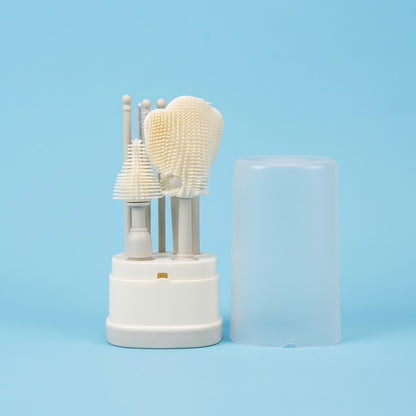 A set of silicone cleaning brushes with beige bristle heads and light gray handles, displayed in a white cylindrical stand next to a translucent clear protective cap on a light blue background.