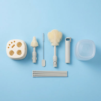 A set of light beige and white baby feeding and cleaning accessories arranged neatly on a solid light blue background. The set includes a round nipple holder, a textured bottle brush, a small cleaning brush, a transfer spoon, a small container, and several transfer tubes.