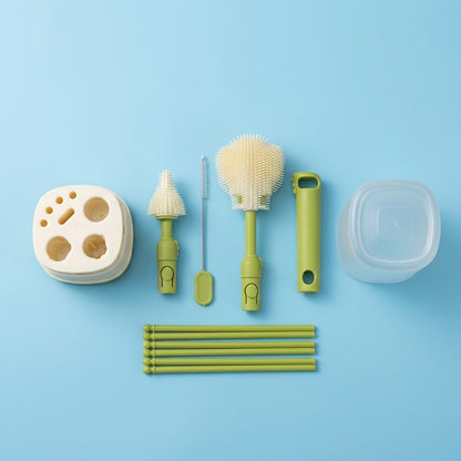 A 6-piece baby bottle cleaning and storage set arranged neatly on a light blue background. The set includes a beige silicone bottle brush with three holes, a small cone-shaped brush, a metal cleaning brush with a green handle, a large round silicone brush, a clear plastic storage container, and six green transfer tubes.