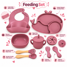 Pink baby feeding set arranged on a white background with callout labels and icons, including a whale-shaped divided plate, silicone suction bowl, sippy cup with straw, wooden-handled baby fork and spoon, silicone baby fork and spoon, and a silicone snack cup lid.