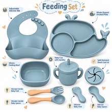 A baby feeding set displayed on a white background with callout labels. The set includes a silicone bib, whale-shaped divided plate, feeding bowl, sippy cup with handles, spill-resistant snack cup lid, silicone baby fork, wooden handle baby spoon, and wooden handle baby fork.