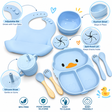A baby feeding set in light blue silicone, including a bib, suction bowl, spill-proof lid, sippy cup with silicone straw, silicone spoon, silicone fork, and a duck-shaped divided plate. The image features callout circles with text labels explaining product features like adjustable bib, suction bowl, spill-proof lid, and silicone straw, along with icons indicating BPA free, food grade silicone, easy to clean, and baby friendly.