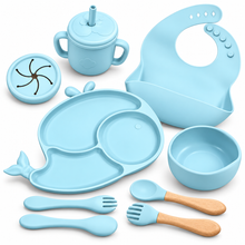 A light blue silicone baby feeding set arranged on a white background, including a divided whale-shaped plate, bowl, bib, sippy cup with lid, two spoons, and two utensils with wooden handles.